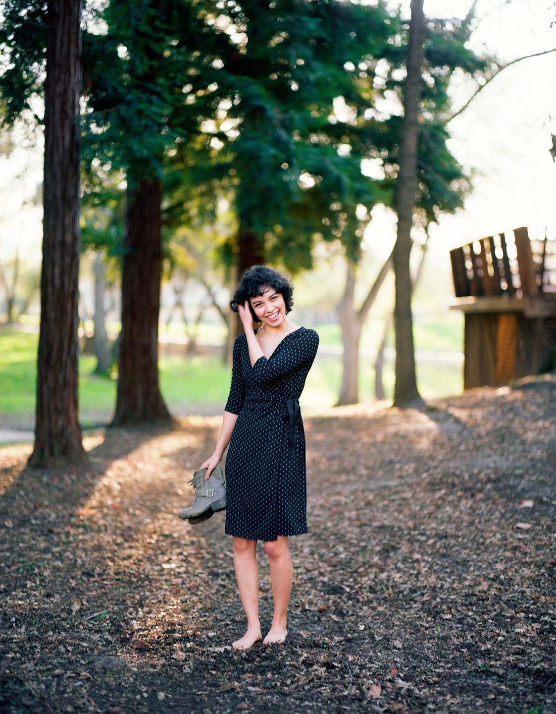 A barefoot woman in a black dress stands on a forest floor covered in leaves, holding a boot in one hand.