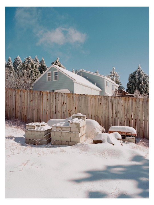 A wooden fence separates a snow-covered yard with concrete blocks from a house with snow on its roof and trees in the background.