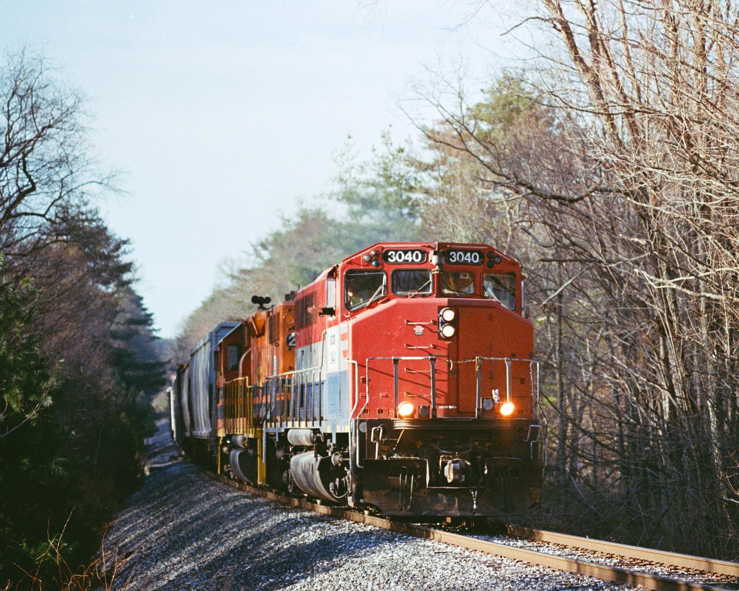 A red and orange train with its headlights on travels along a track through a forest of bare trees.