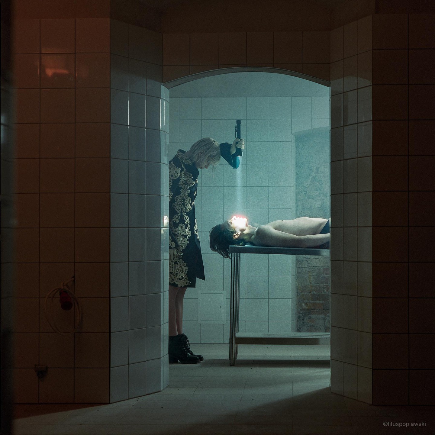 A woman in a patterned coat shines a light on a man lying on a metal table in a tiled room.