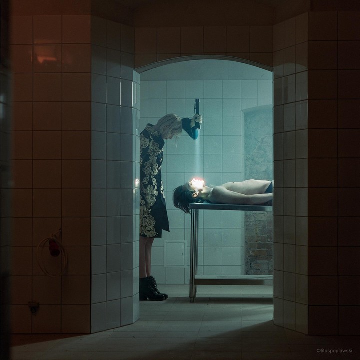 A woman in a patterned coat shines a light on a man lying on a metal table in a tiled room.