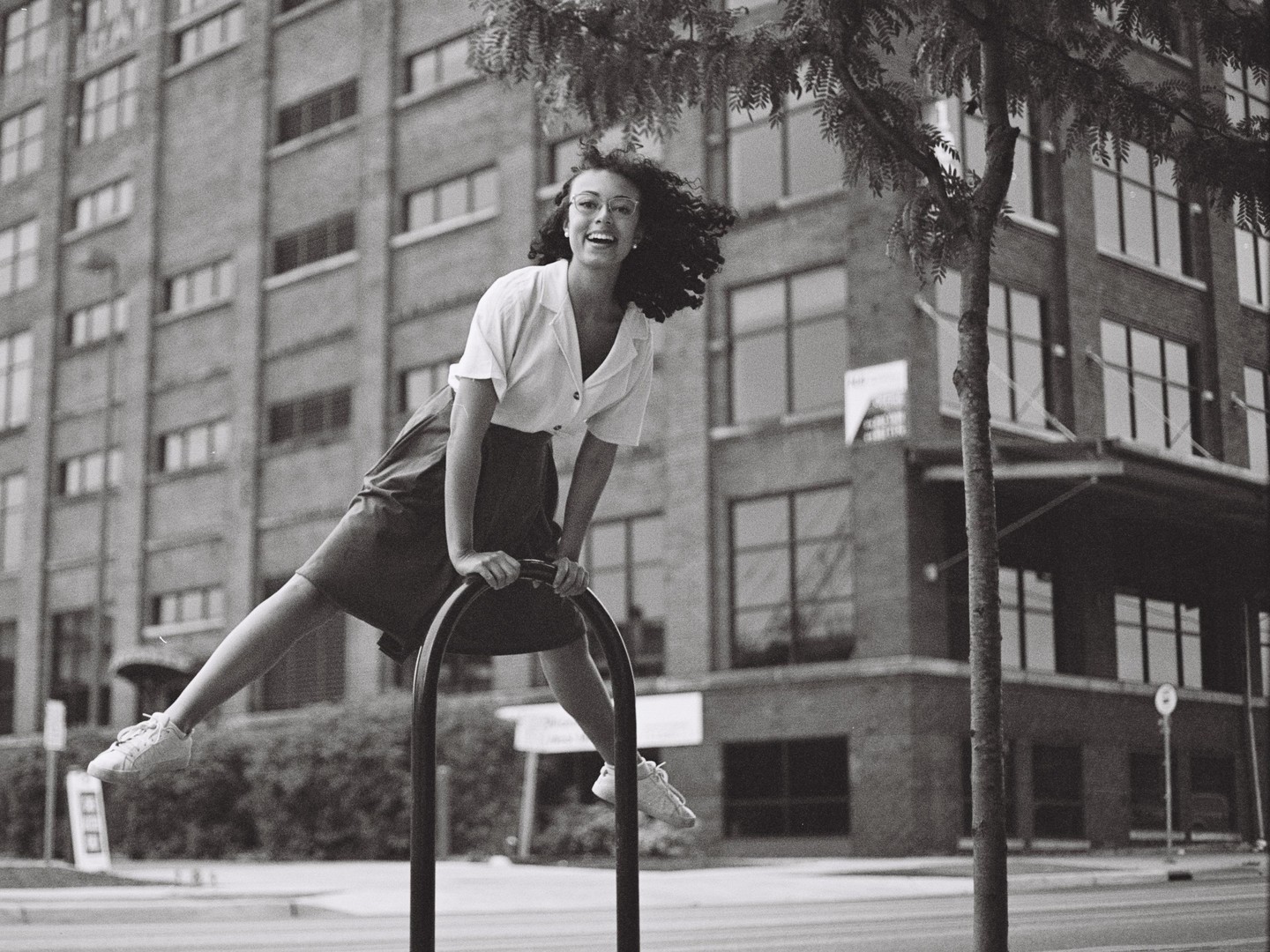 A woman in a skirt and shirt is balancing on a bike rack in front of a building.
