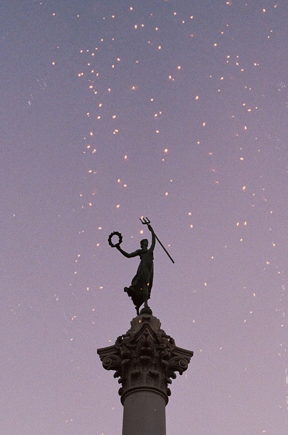 A statue of a woman holding a trident and a wreath stands atop a column against a purple sky with sparkling lights.