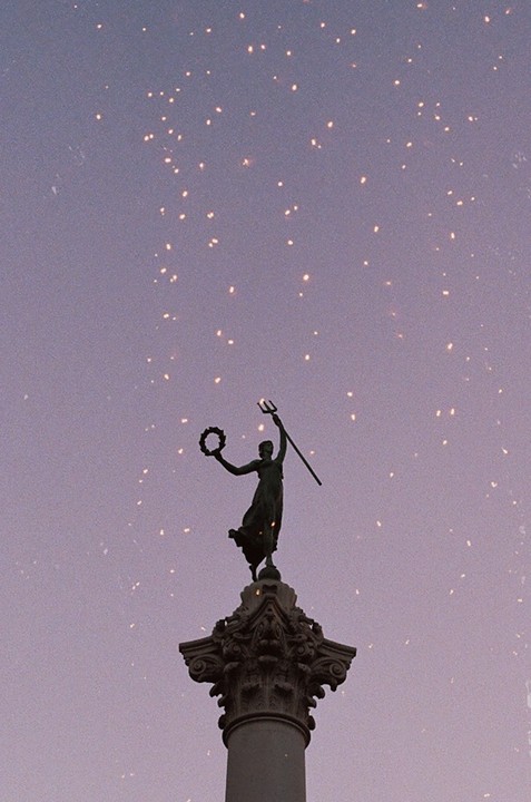 A statue of a woman holding a trident and a wreath stands atop a column against a purple sky with sparkling lights.