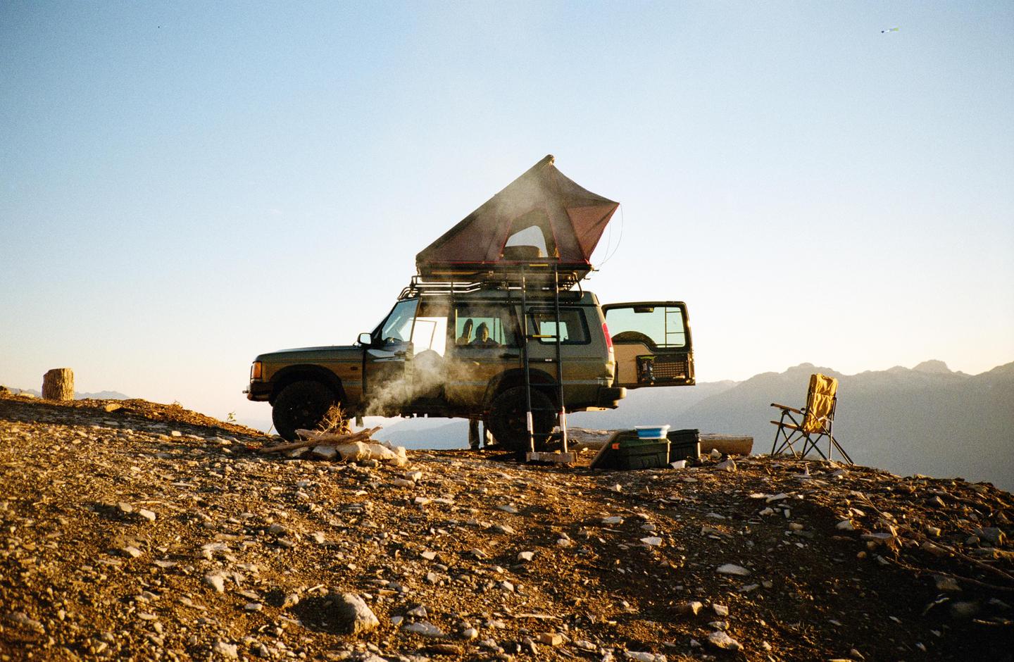 A vehicle with a rooftop tent is parked on a rocky hill with mountains in the background.