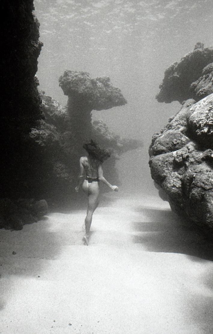 A woman in a bikini walks along a sandy underwater path between large rock formations.