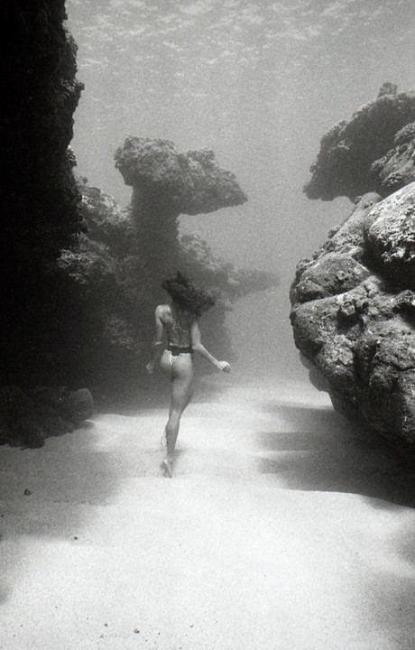 A woman in a bikini walks along a sandy underwater path between large rock formations.