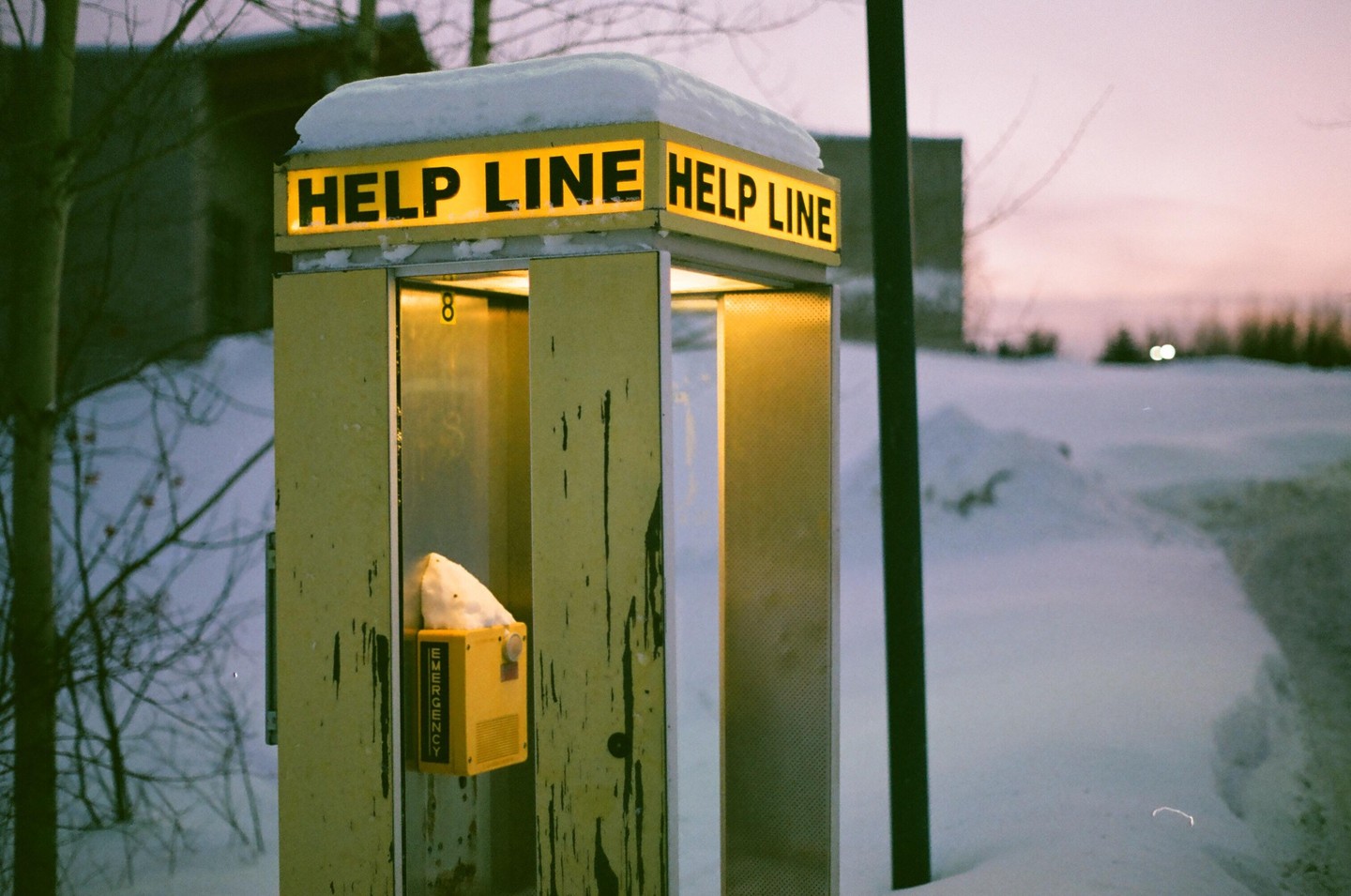 A yellow help line booth with snow on its roof stands in a snowy landscape under a pink sky.