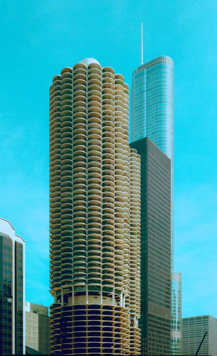 A tall, cylindrical building with many balconies stands next to other skyscrapers under a bright blue sky.