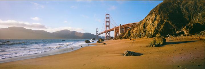 The Golden Gate Bridge is visible from a sandy beach with waves crashing on the shore and a large cliff on the right.