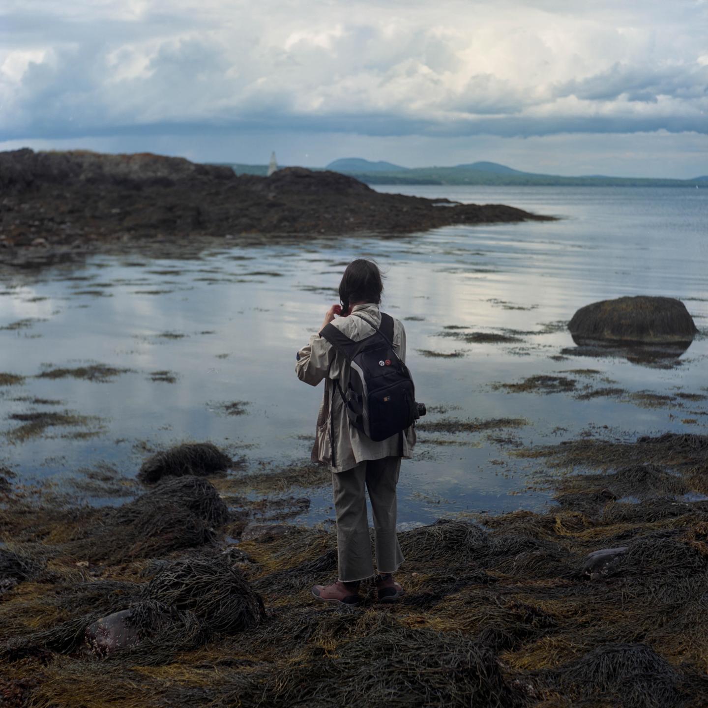 A woman with a backpack stands on a rocky, seaweed-covered shore looking out at the water and distant mountains under a cloudy sky.