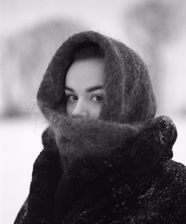 A woman wearing a dark, fuzzy hood and coat looks directly at the viewer, with snow visible on her clothing.