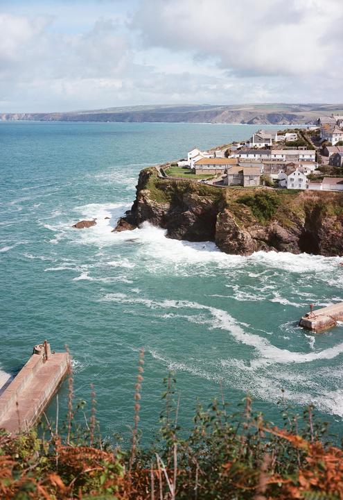 A coastal village with white houses sits atop a cliff overlooking a turquoise sea with crashing waves and a small harbor.