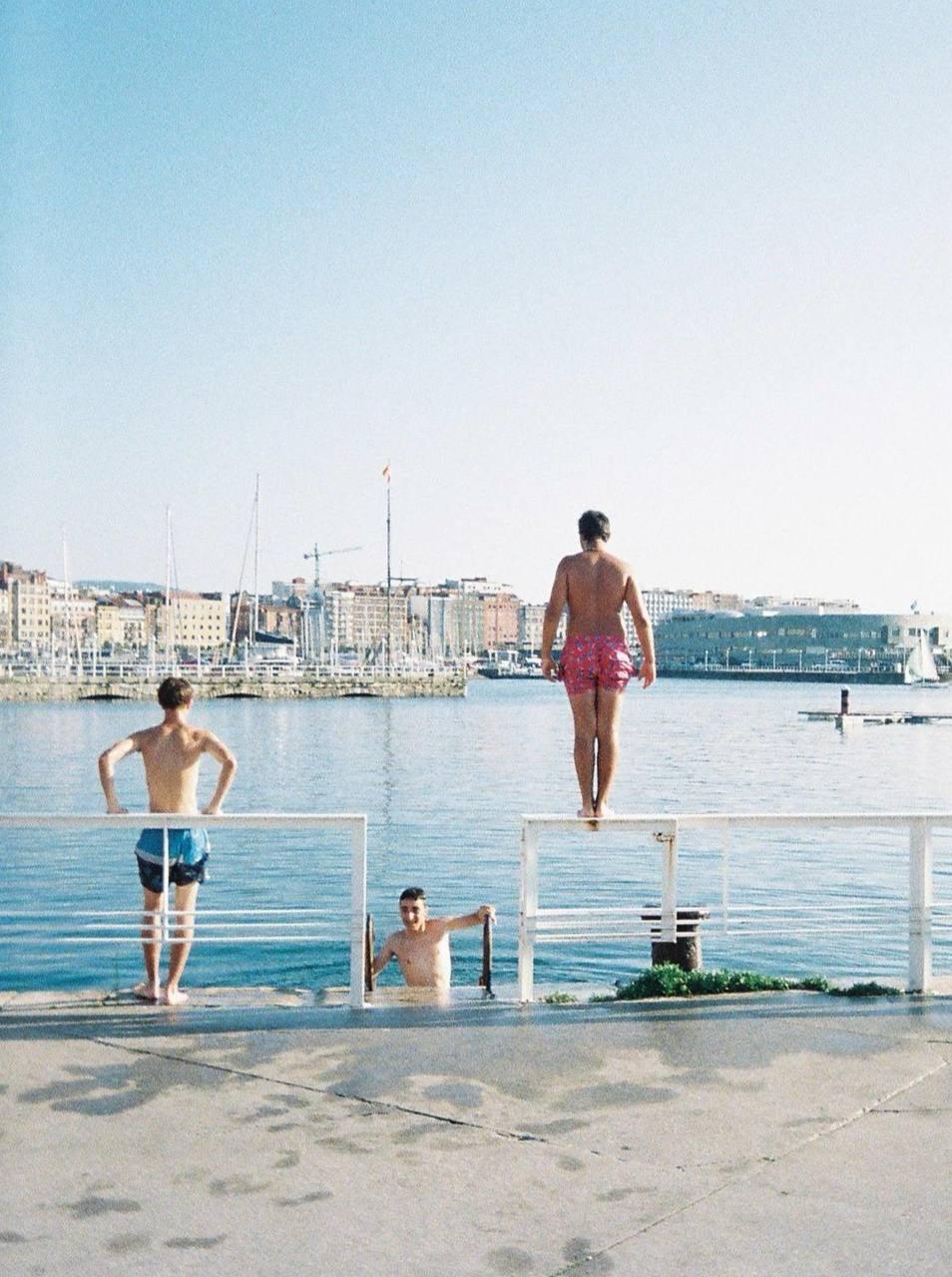 Three men are at a pier, with two standing on the pier and one in the water, overlooking a harbor with boats and buildings.