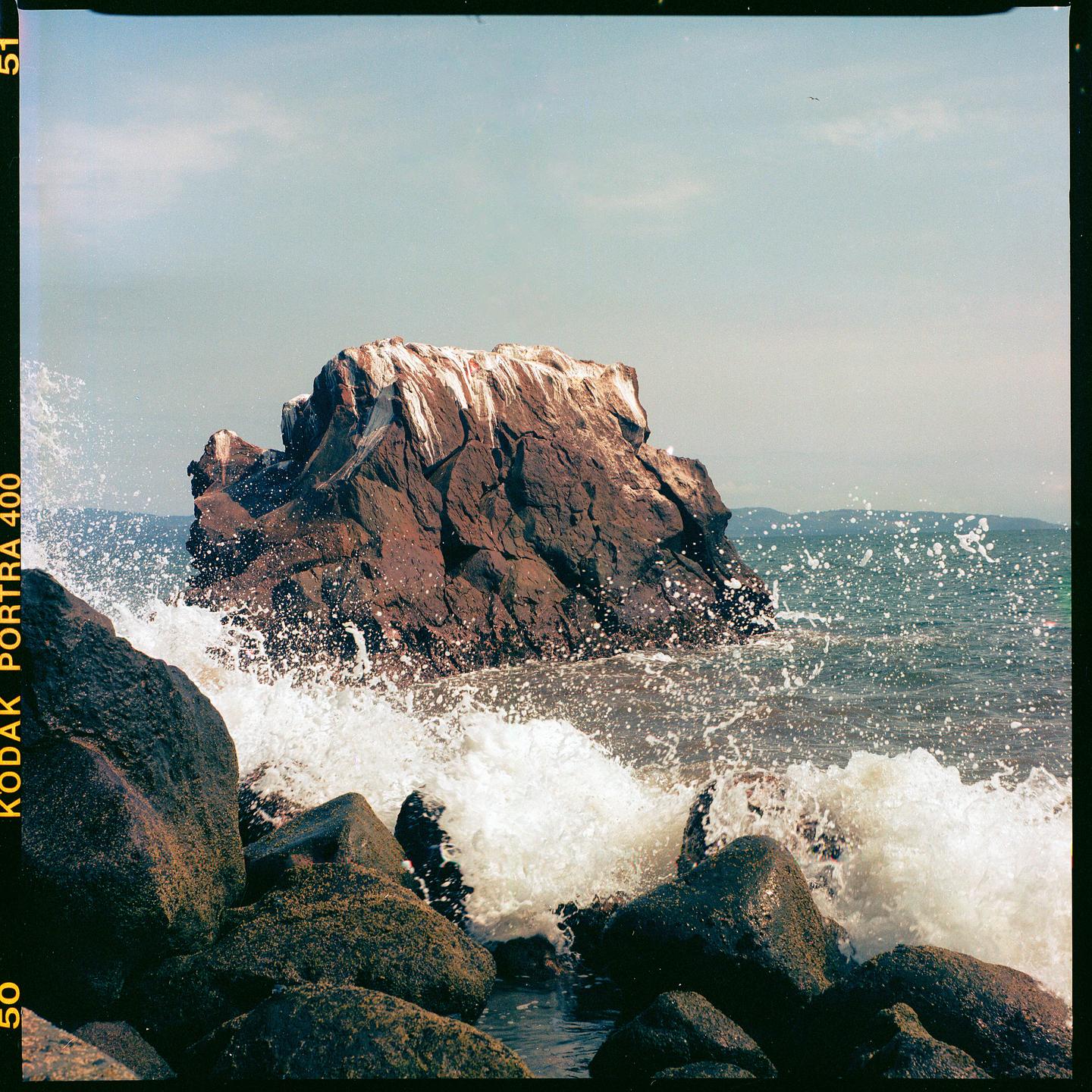 Waves crash against rocks on a coastline with a large rock formation in the distance.
