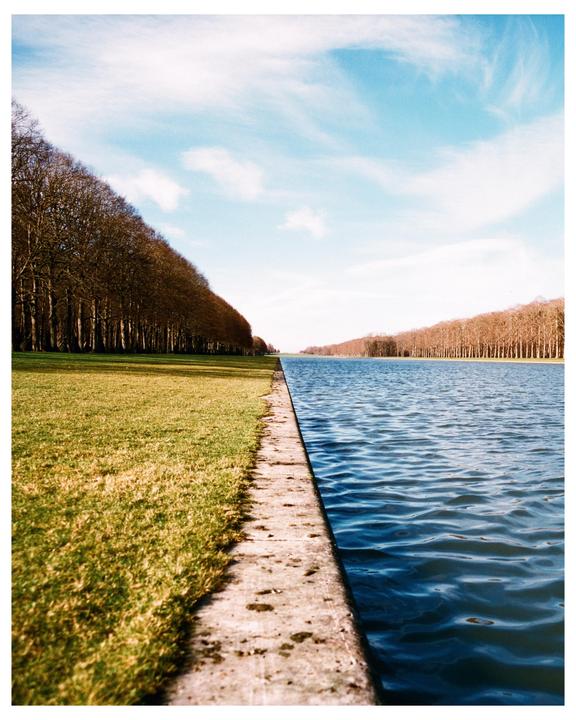 A long canal with trees lining both sides stretches into the distance under a blue sky with wispy clouds.