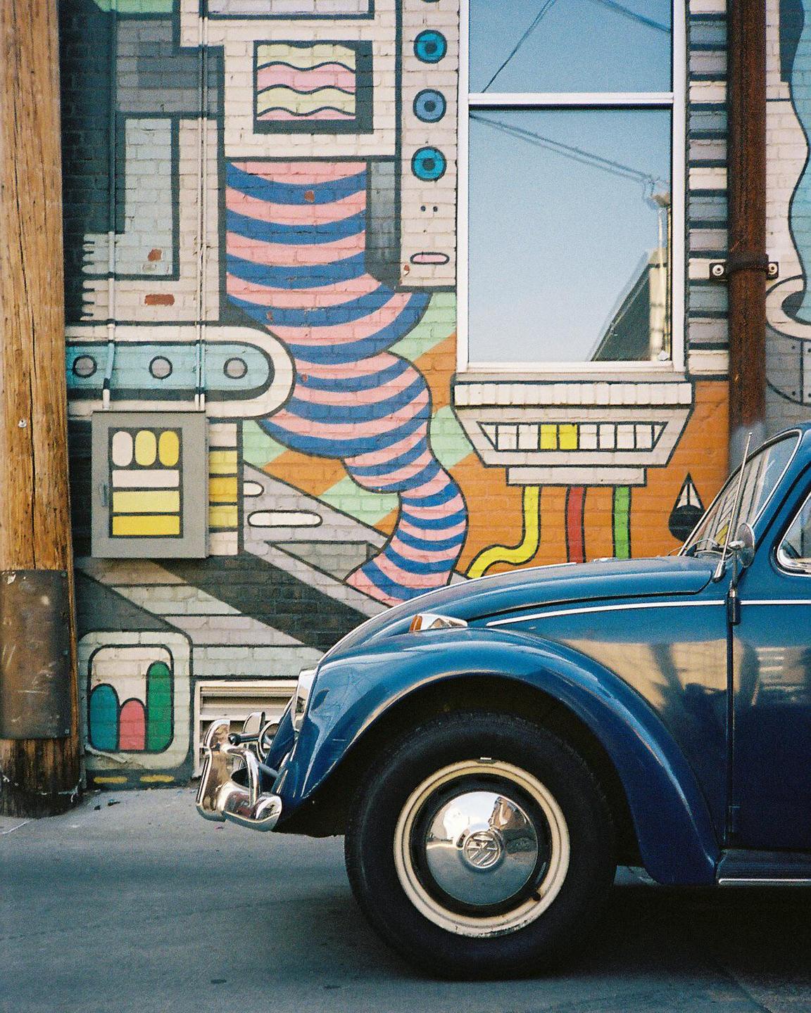 A blue car is parked next to a building with a colorful mural and a window.