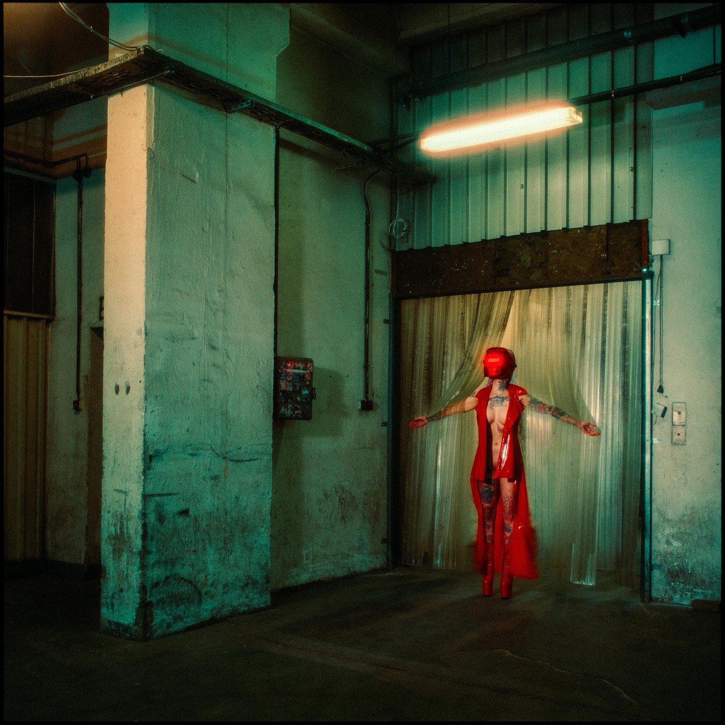 A woman in a red outfit and helmet stands with outstretched arms in a dimly lit industrial space.