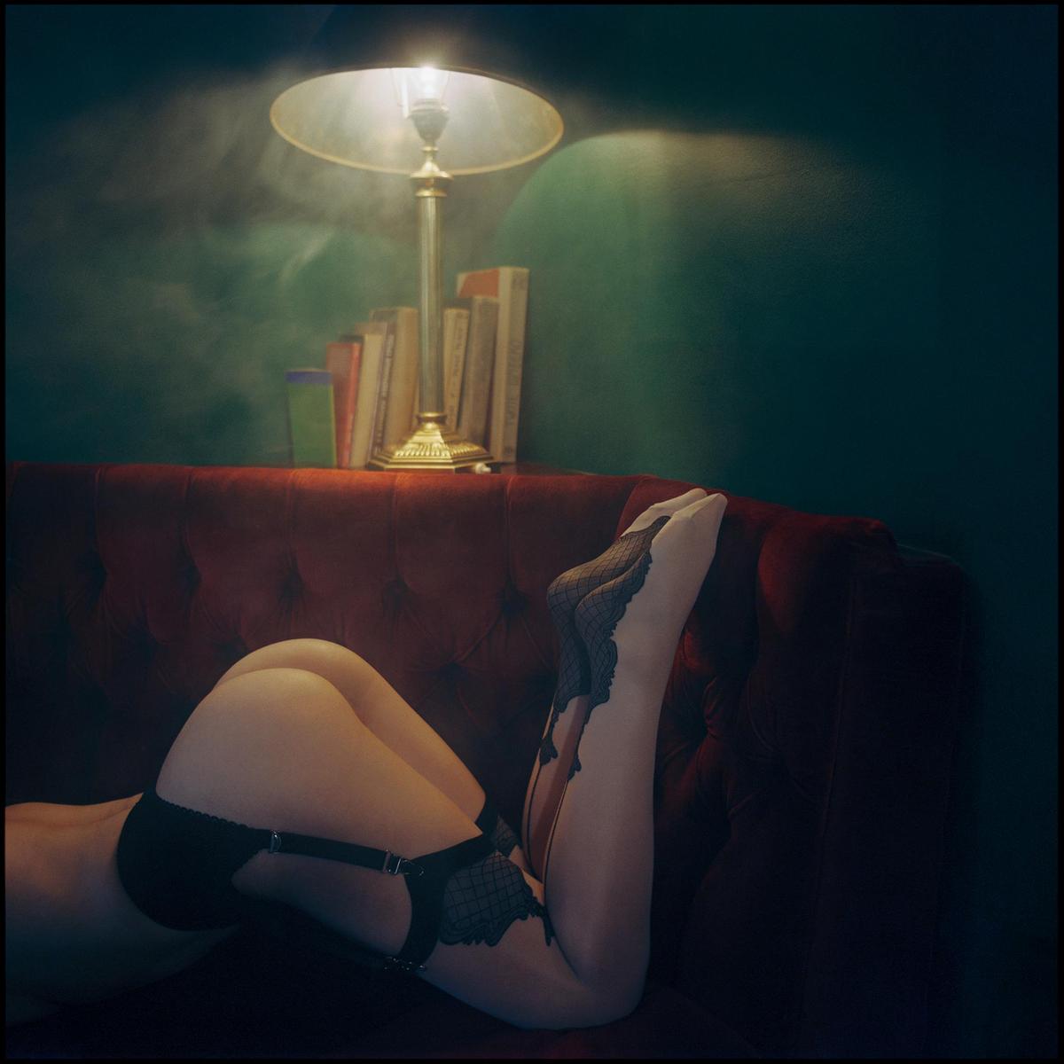 A woman in lingerie is lying on a red sofa with a lamp and books in the background.