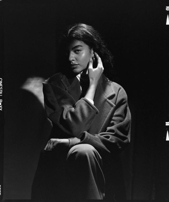 A woman in a coat sits with her hand near her face, with part of her face in shadow.