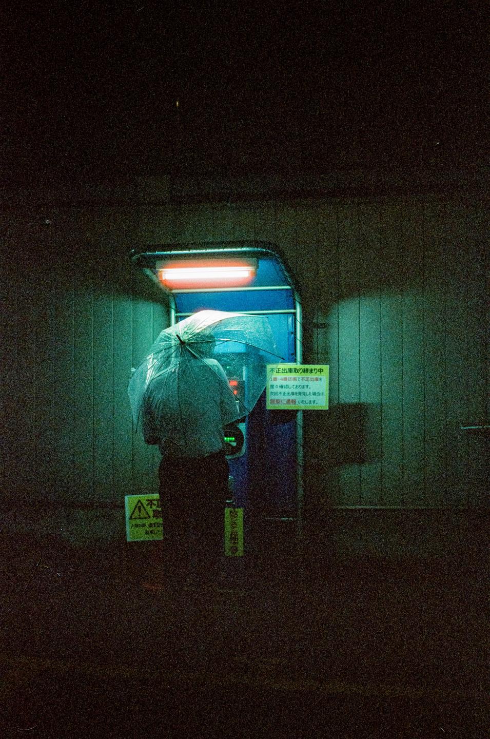 A person holding an umbrella stands in front of an ATM at night in Hiroshima.