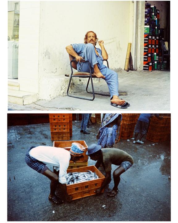 A man with a mustache sits on a chair next to a building, and two men carry a crate of fish on a wet street.