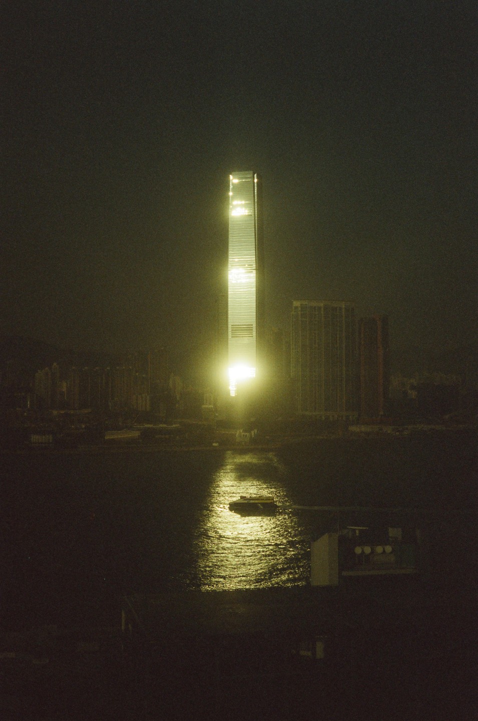 A tall, illuminated skyscraper reflects its light onto the water below, where a small boat floats.