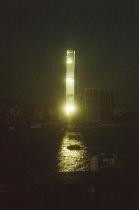 A tall, illuminated skyscraper reflects its light onto the water below, where a small boat floats.