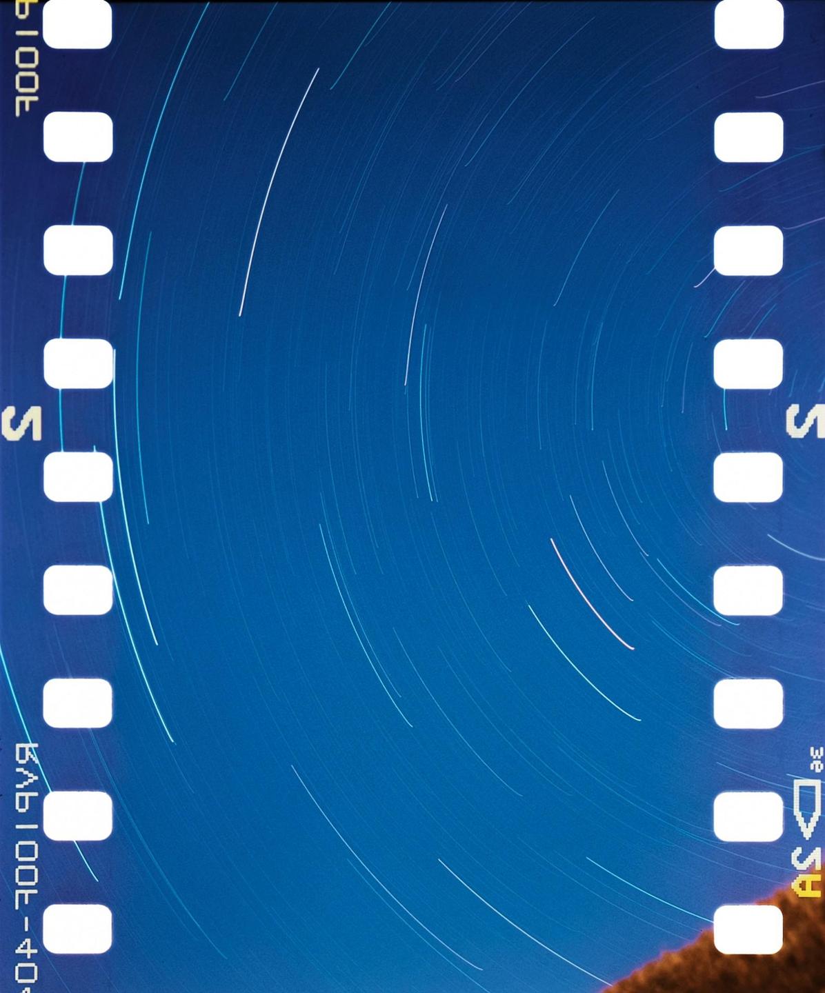 A blue night sky with star trails is visible through the sprocket holes of a film strip.