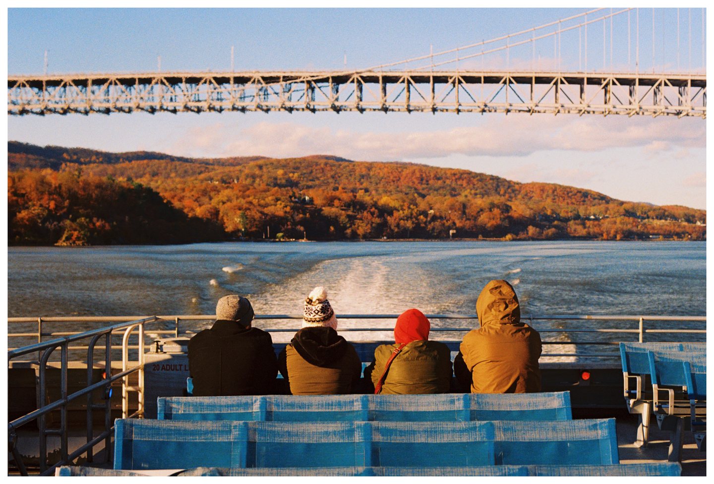 Four people sit on a ferry looking at a bridge and a tree-covered hill with autumn foliage.