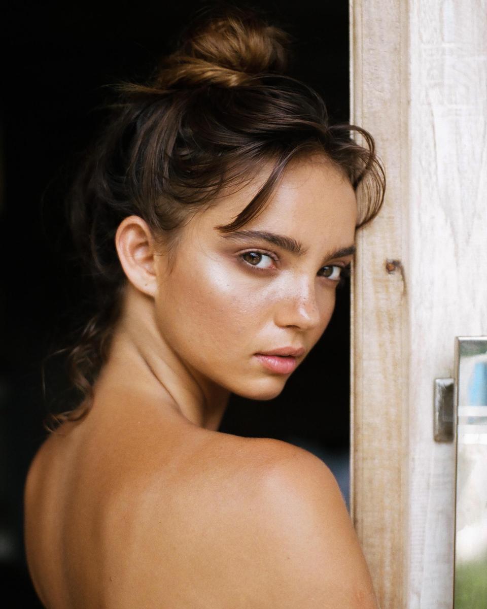 A woman with her hair in a bun looks over her shoulder while leaning against a wooden doorframe.
