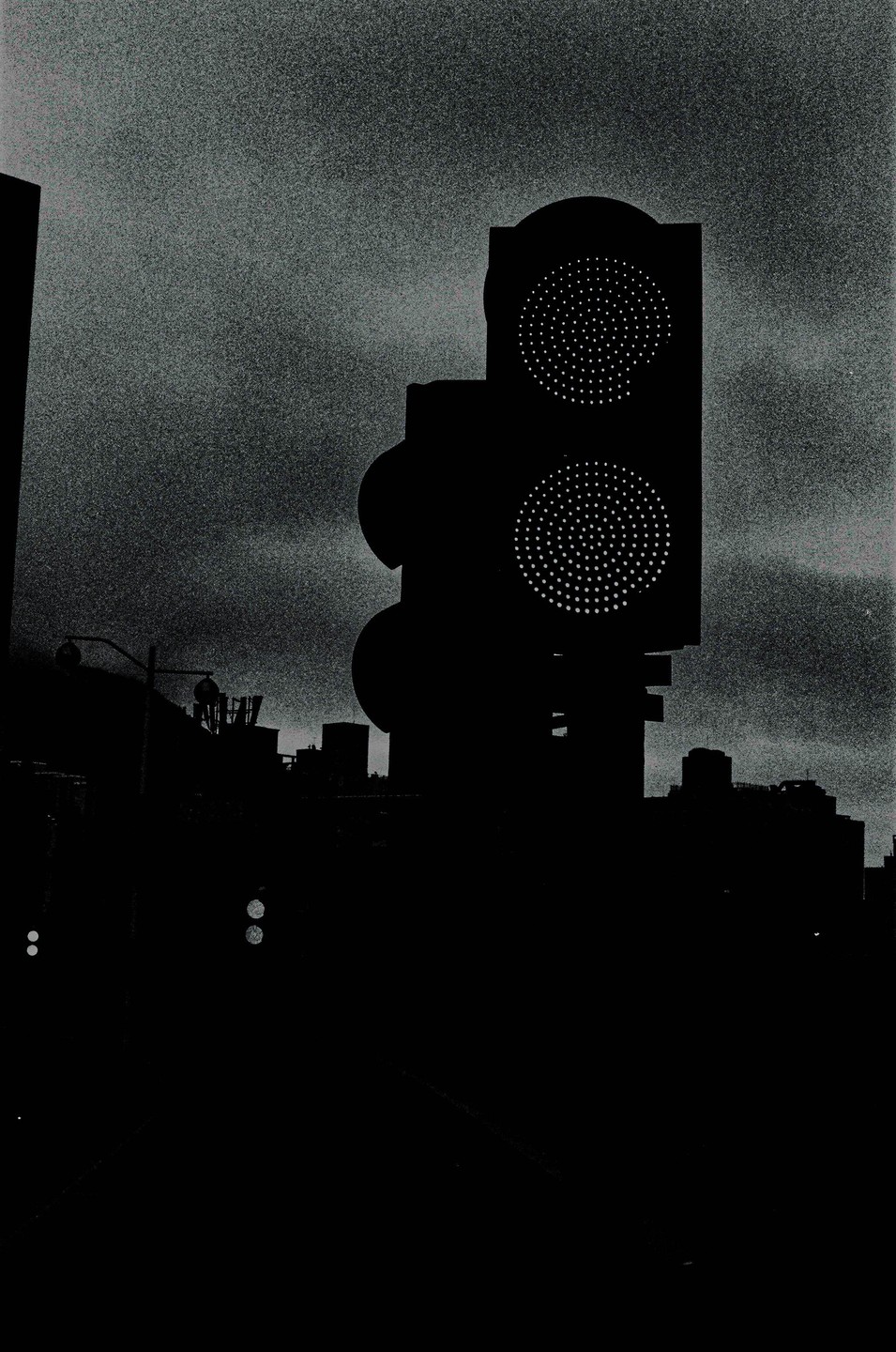 A traffic light with two illuminated signals stands against a dark, grainy sky above a city skyline.