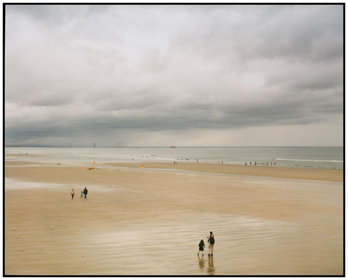 A wide sandy beach with people walking along the shore under a cloudy sky.