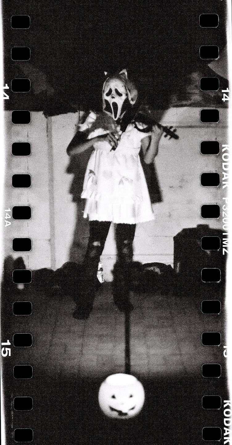 A person in a ghostface mask and white dress plays a violin while standing over a pumpkin pail.