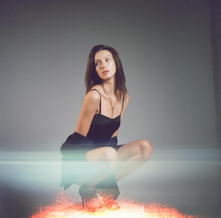 A woman in a black dress and high heels crouches on a reflective surface with a light leak and red light.