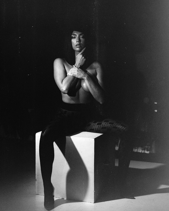 A woman sits on a white cube, her arms crossed over her chest, with a spotlight creating a strong shadow behind her.