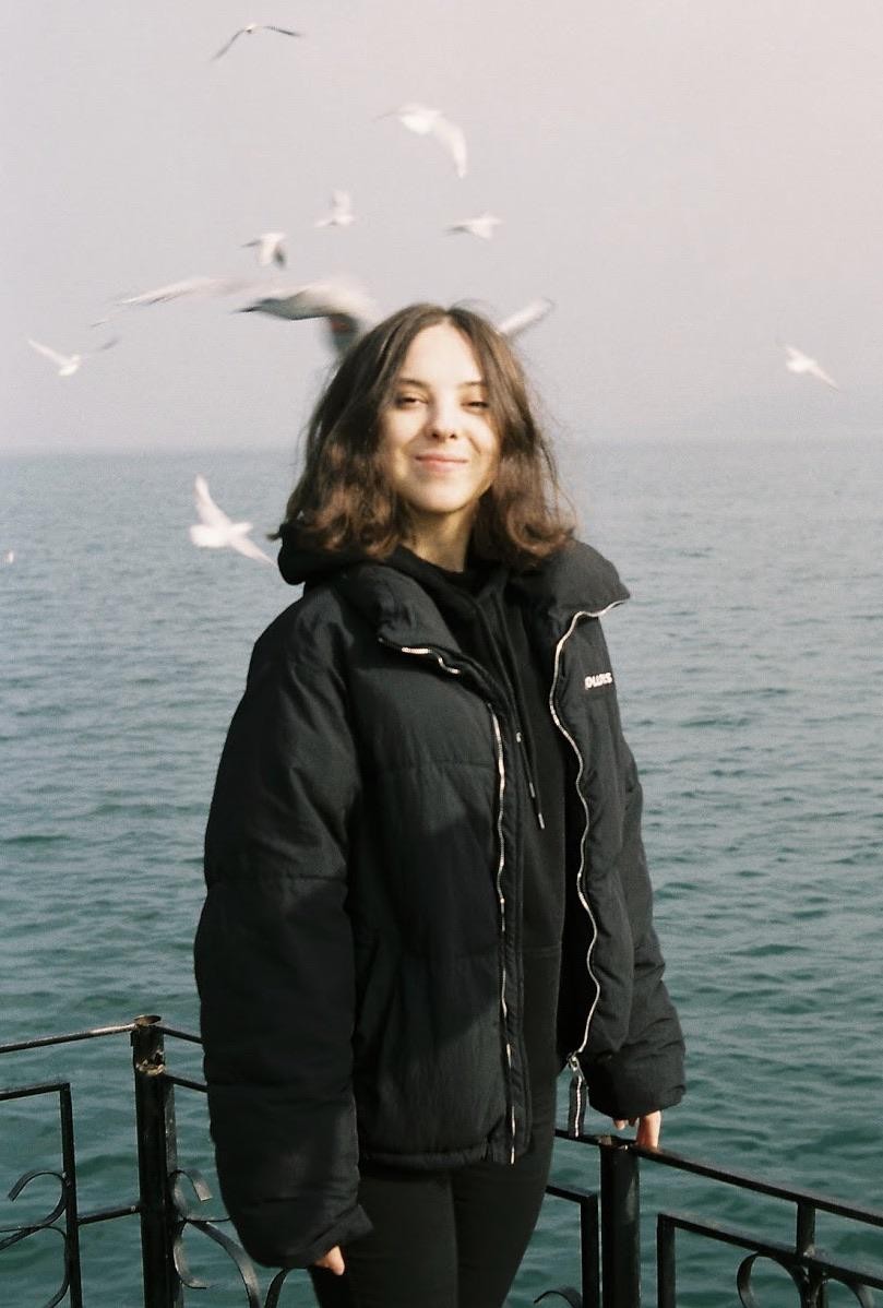A woman in a black jacket stands by the water with seagulls flying in the background.