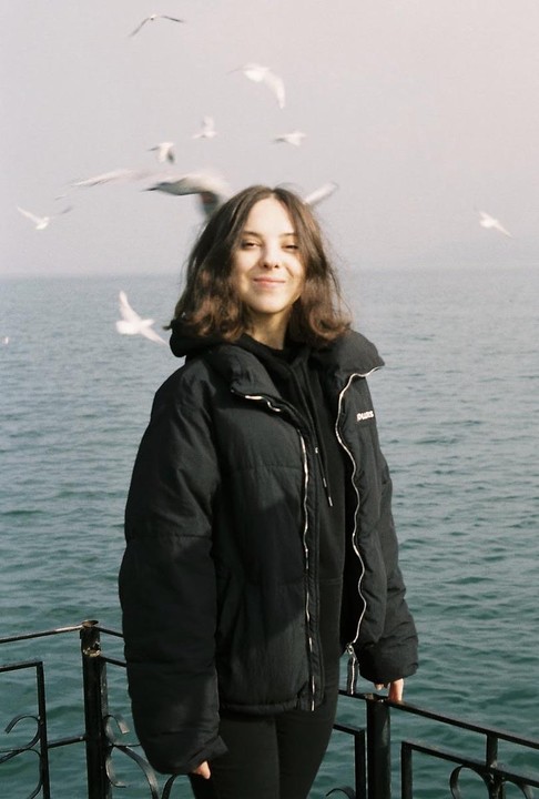 A woman in a black jacket stands by the water with seagulls flying in the background.