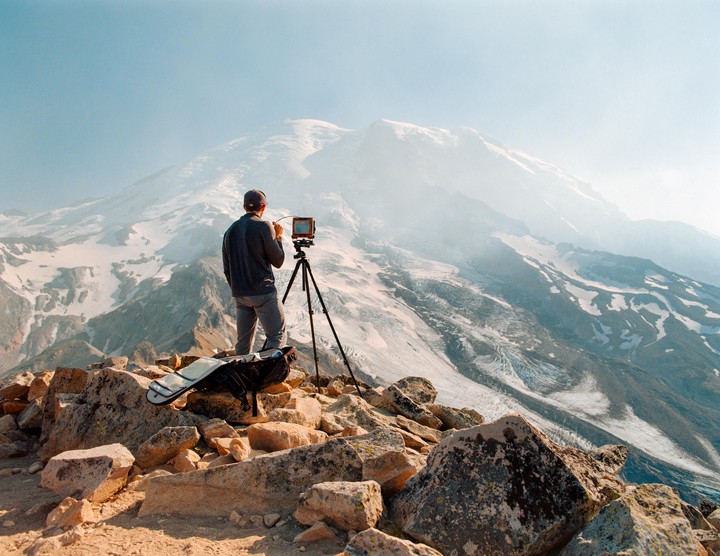A man stands on a rocky mountain peak, operating a camera on a tripod, with a vast snowy mountain range in the background.
