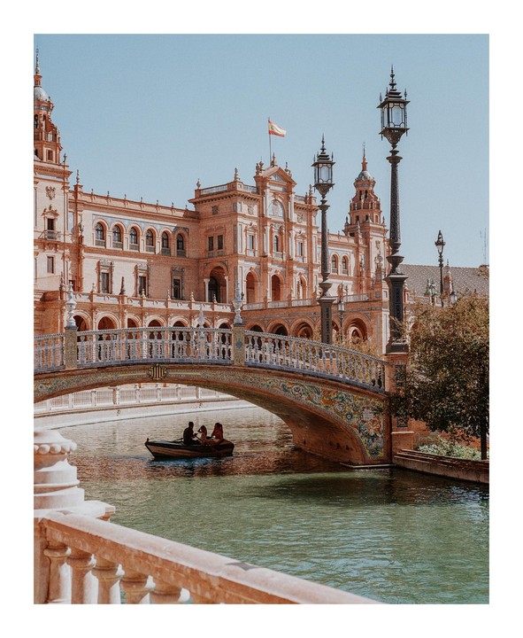 A small boat with three people floats on a canal under a decorative bridge in front of a large, ornate building.