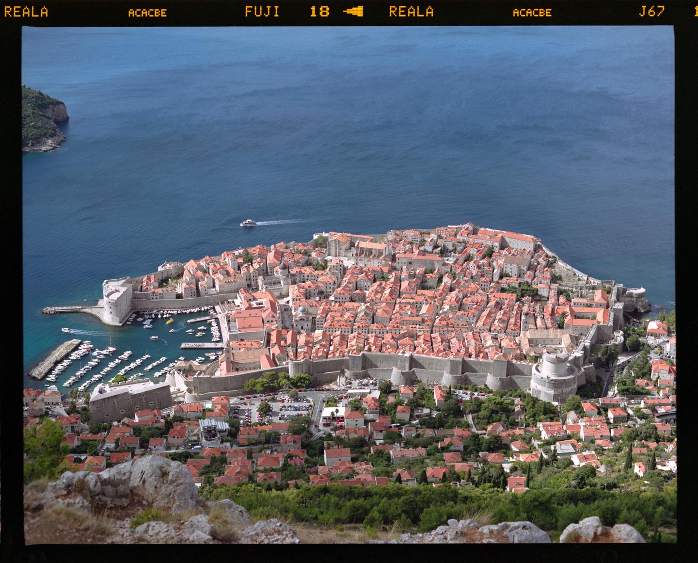 An aerial view shows a walled city with red-tiled roofs nestled beside a harbor with boats, surrounded by blue water and green hills.