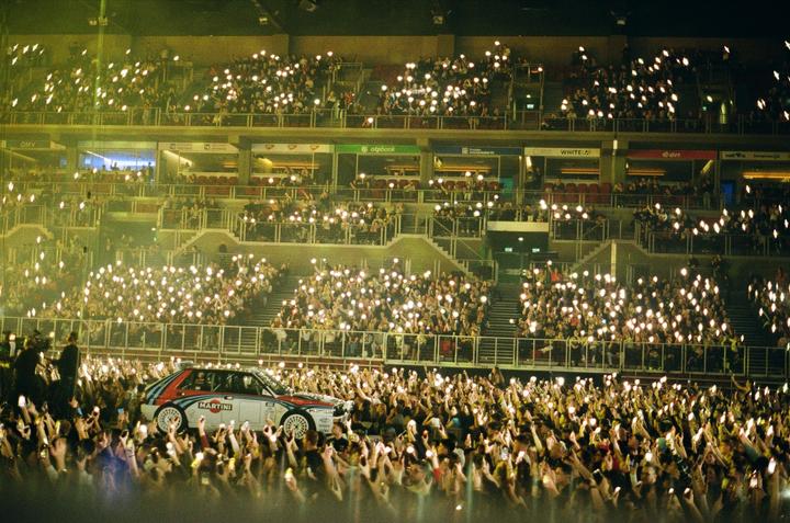 A rally car is surrounded by a crowd of people holding up lights in a stadium at night.