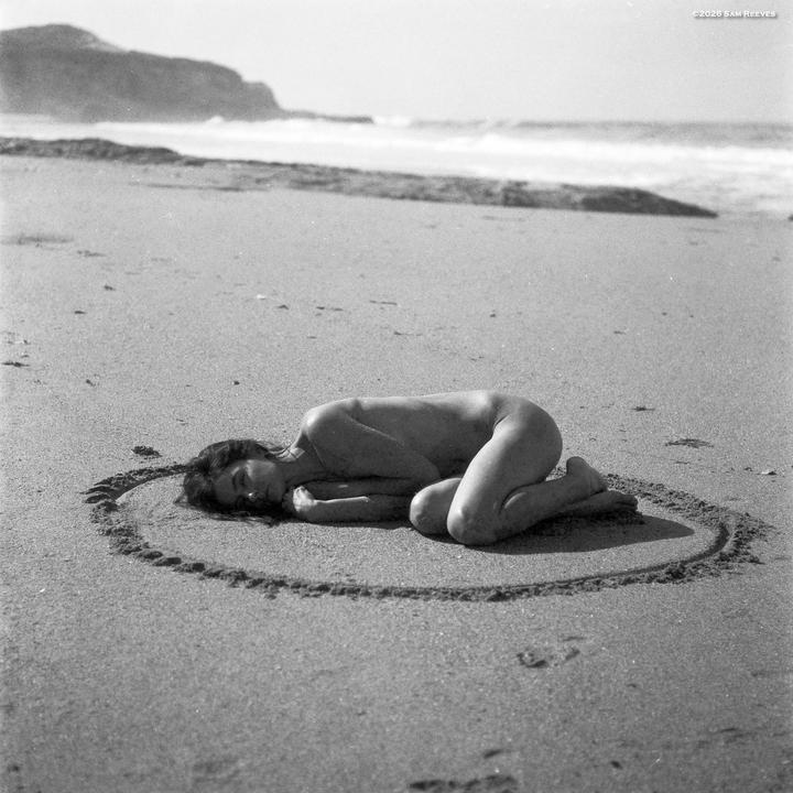 A woman lies nude in a circle drawn in the sand on a beach with waves and a cliff in the background.