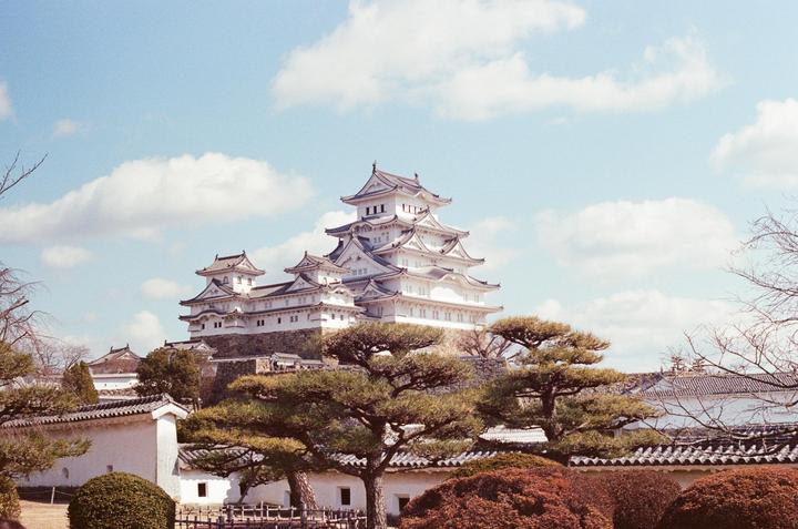 A large white multi-tiered castle with dark roofs stands on a hill surrounded by trees and traditional Japanese buildings under a blue sky with white clouds.