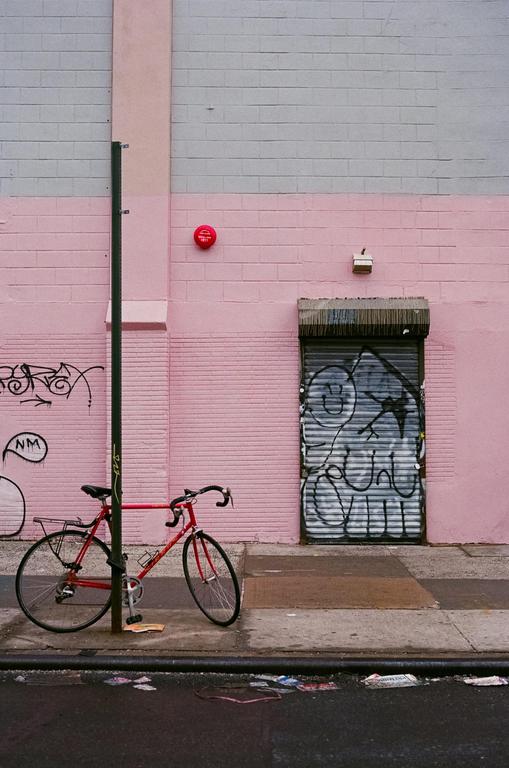 A red bicycle is parked next to a pink and gray building with graffiti on a metal door.