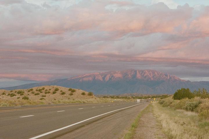 A road stretches towards mountains under a sky filled with pink clouds at sunset.