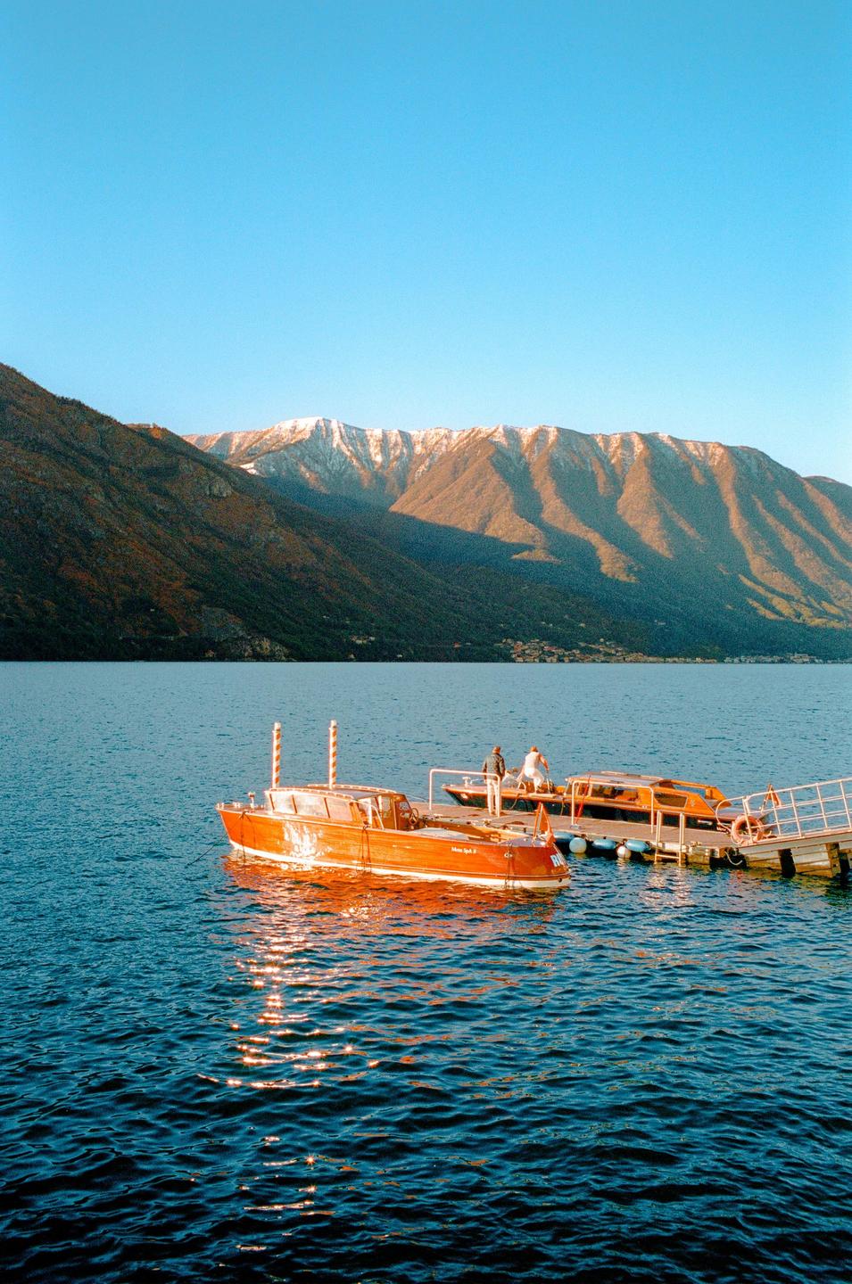 Two boats are docked at a pier on a lake with mountains in the background.