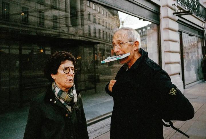 A man holds a small book in his mouth while a woman looks at him on a city street.