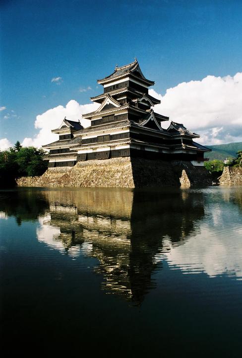 A traditional Japanese castle with multiple dark tiers and white trim is reflected in the still water of a moat under a blue sky with white clouds.