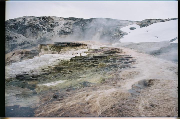 Steam rises from terraced hot springs with snow-covered mountains in the background.